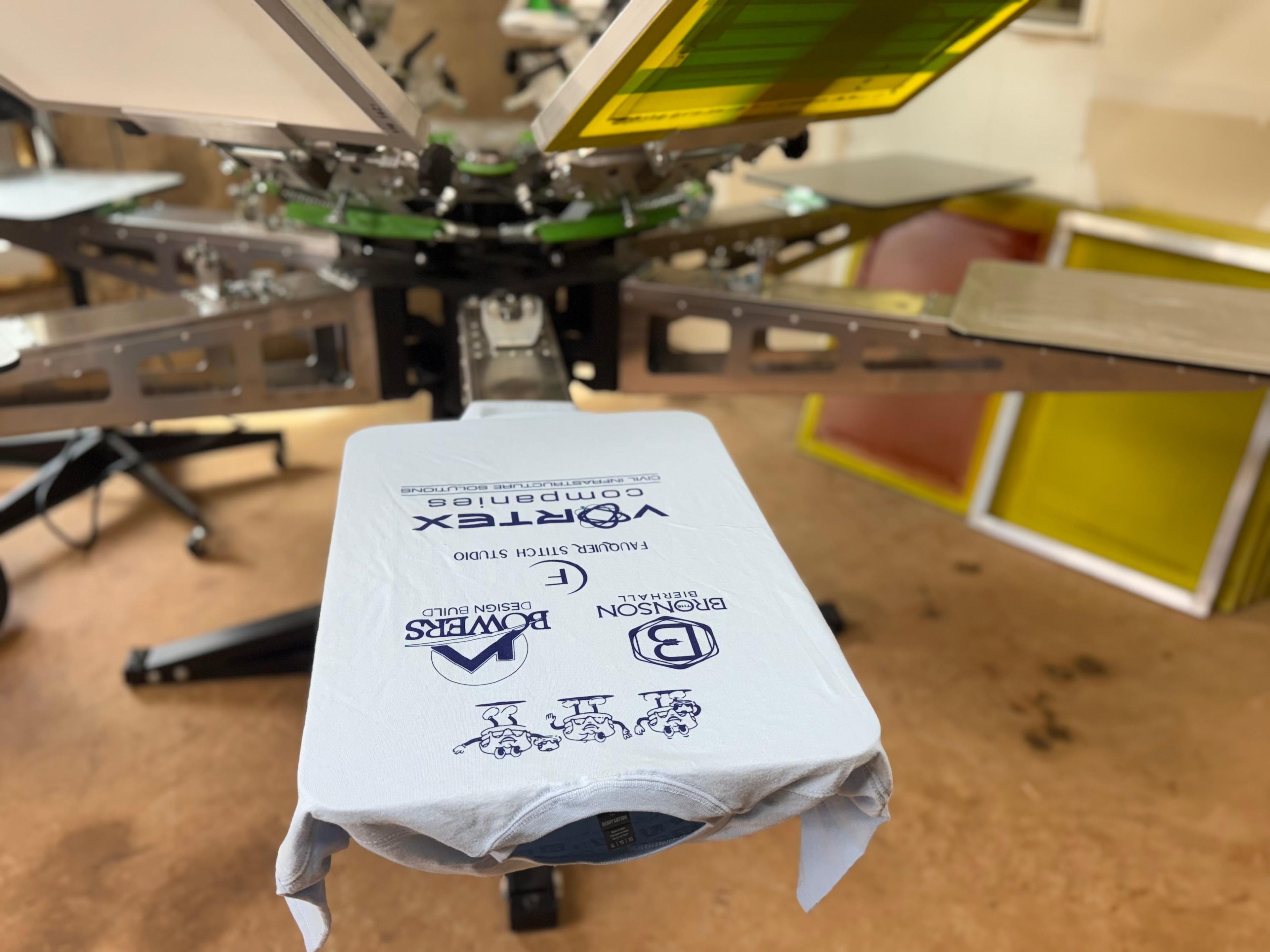Screen printing machine setup with a white T-shirt displaying blue text and logos, ready for printing.