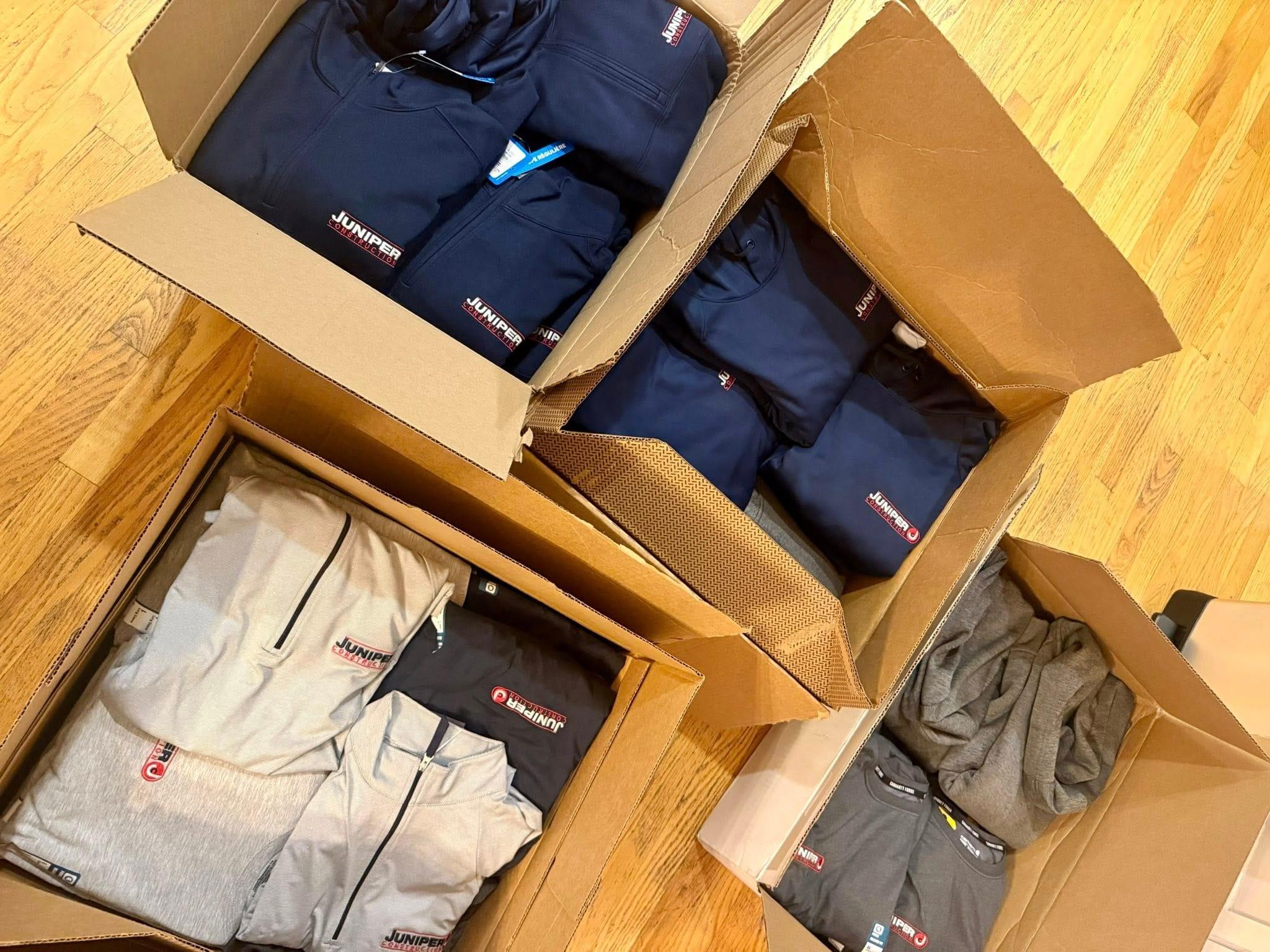 Cardboard boxes on a wooden floor, each filled with neatly folded clothing items featuring a logo that says "Juniper".