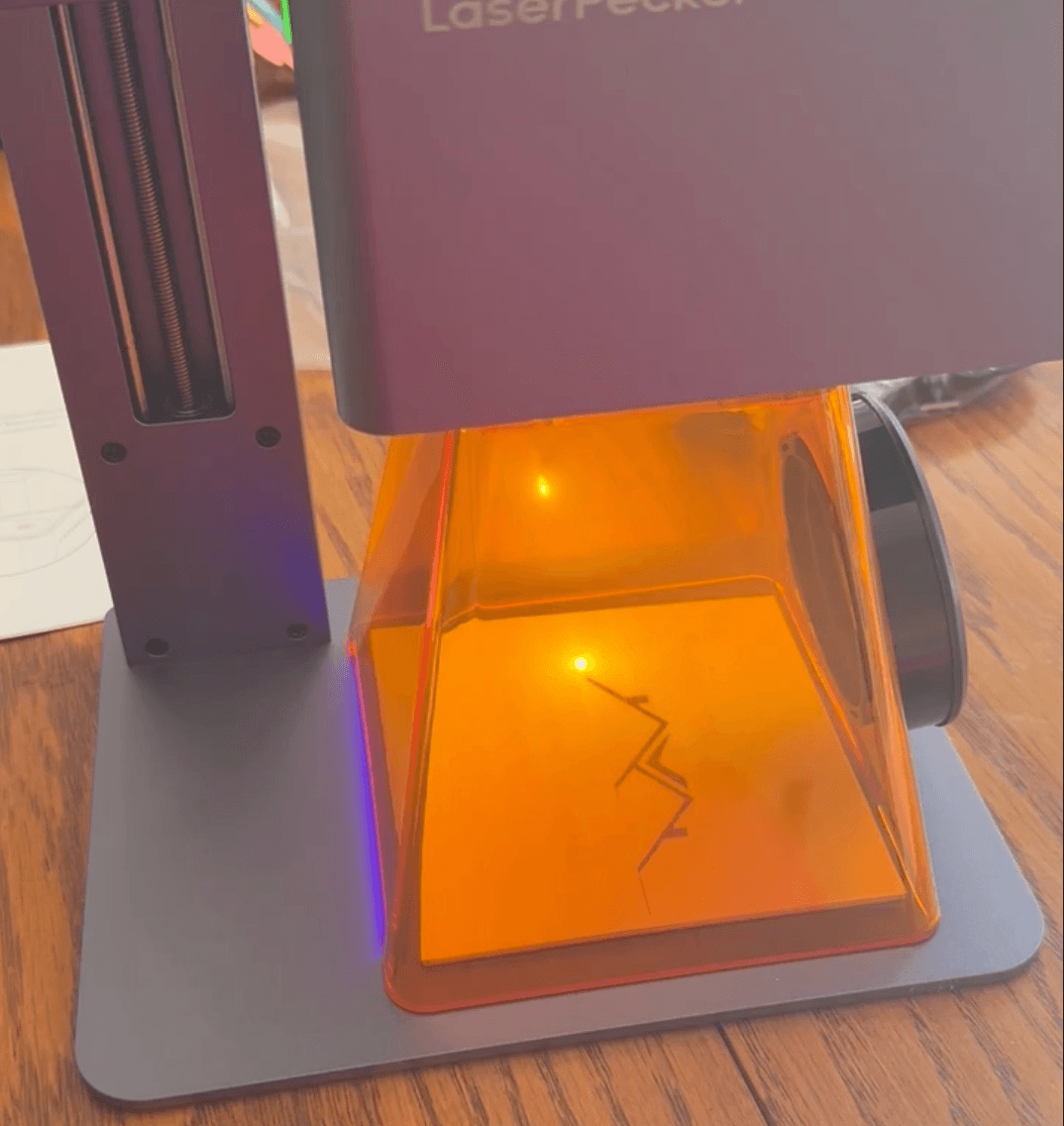 Laser engraver with protective orange cover on a wooden surface, displaying a zigzag pattern being engraved.