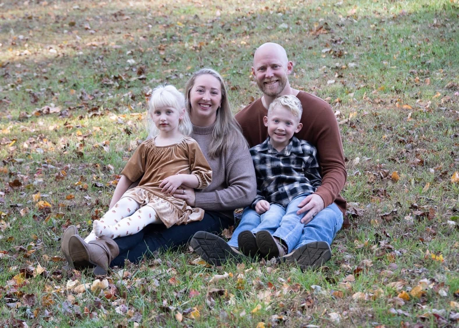 Family sitting on grass in a park, surrounded by autumn leaves, with a woman and man smiling, and two young children dressed in fall attire.