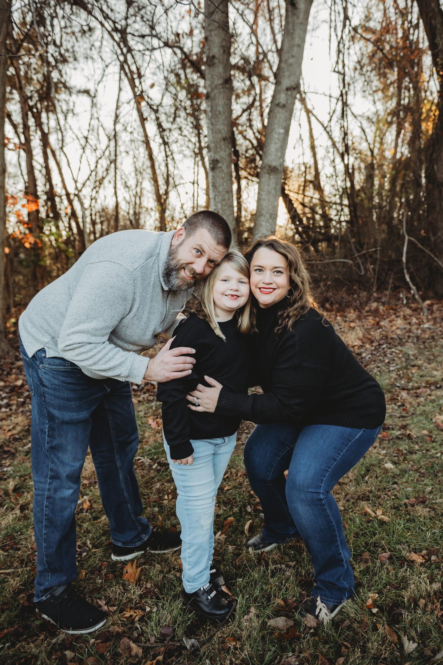 A smiling family of three poses together in a wooded area during fall, with dry leaves scattered on the grass. The father wears a grey sweater, the mother a black top, and the child is dressed in a black shirt and light blue jeans.