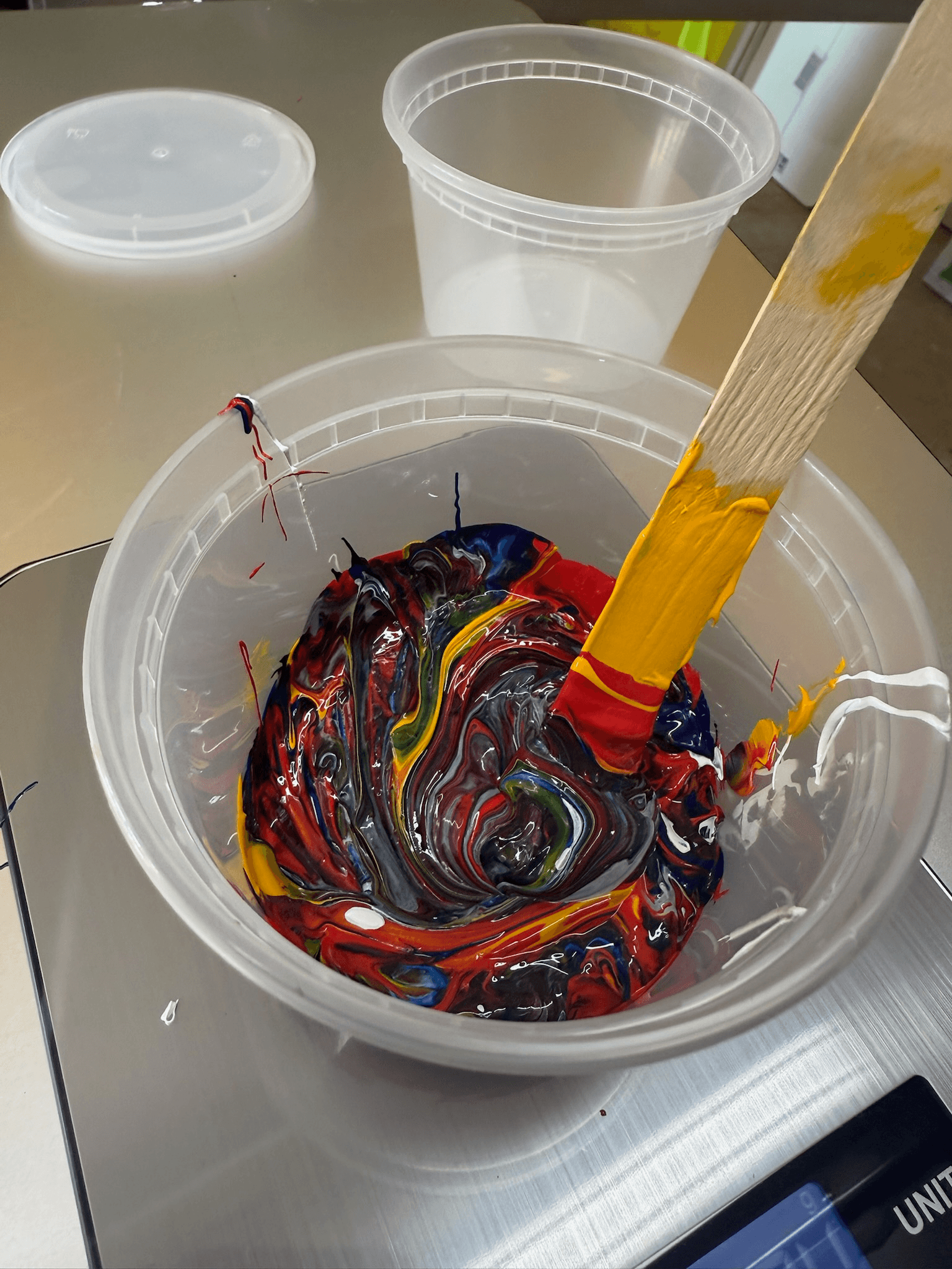 A plastic container filled with a swirling mix of colorful paints, including red, yellow, blue, and black, blended with a wooden stick partially coated in yellow paint. Another empty container and a lid are nearby.