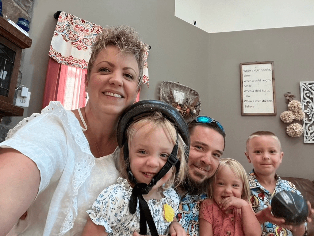 Family of five smiling indoors, with two adults and three children. The young girl in the foreground wears a helmet. The background features decorative signs and floral patterns.