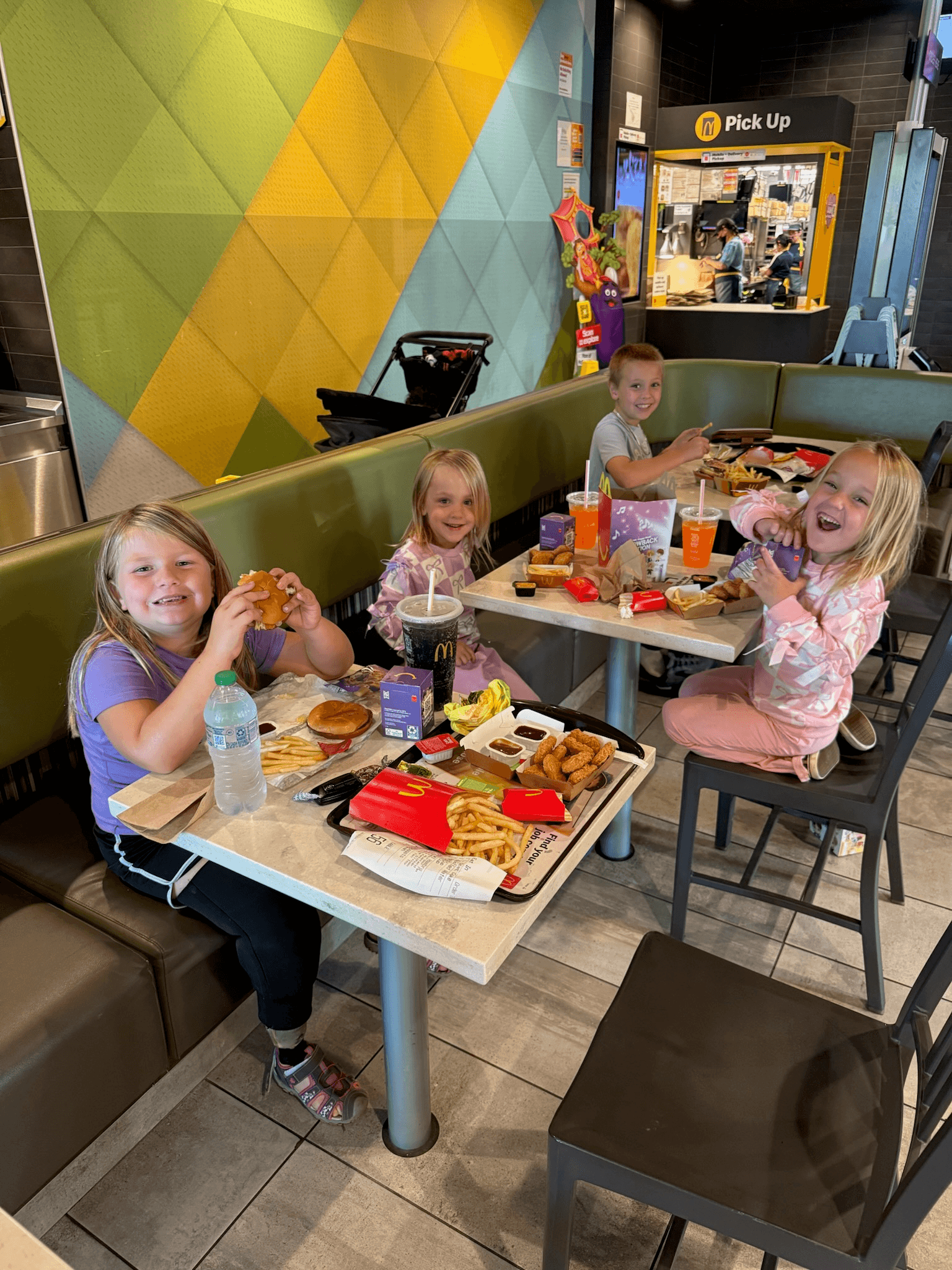 Four children sitting in a fast-food restaurant booth, smiling and eating meals that include fries, chicken nuggets, and drinks. The restaurant features a colorful geometric wall pattern and a visible pick-up counter in the background.