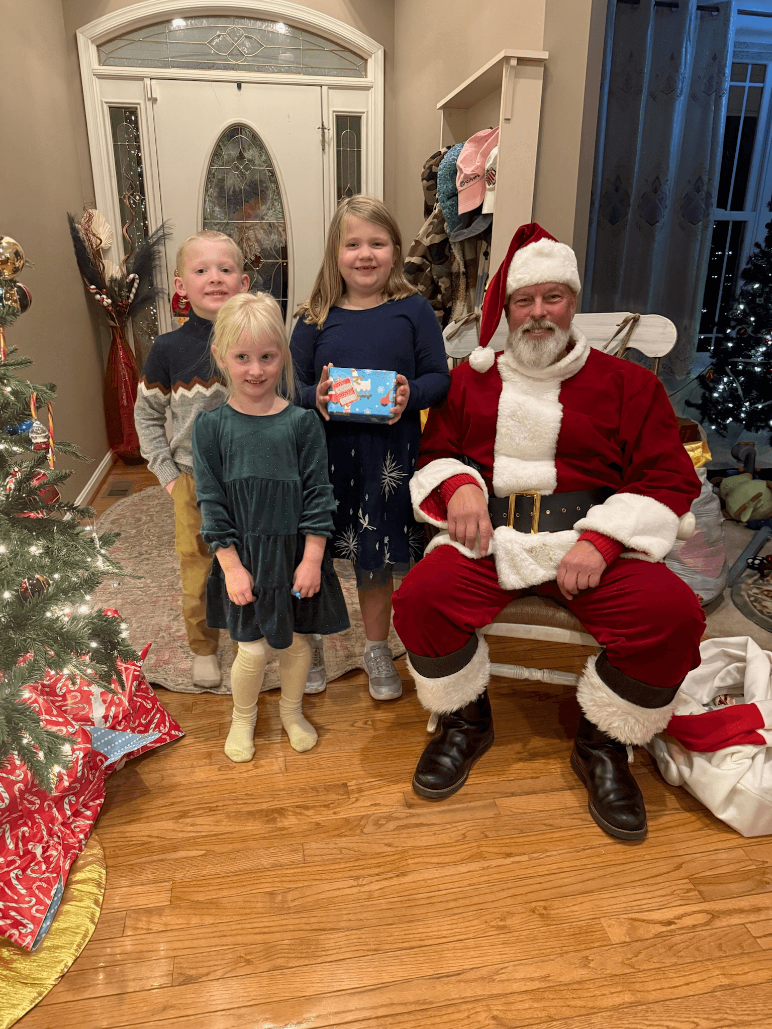 Three children smiling with a man dressed as Santa Claus. The girl in the center holds a wrapped gift. A Christmas tree with decorations and presents is visible on the left, set in a warmly lit room with a decorated entryway.
